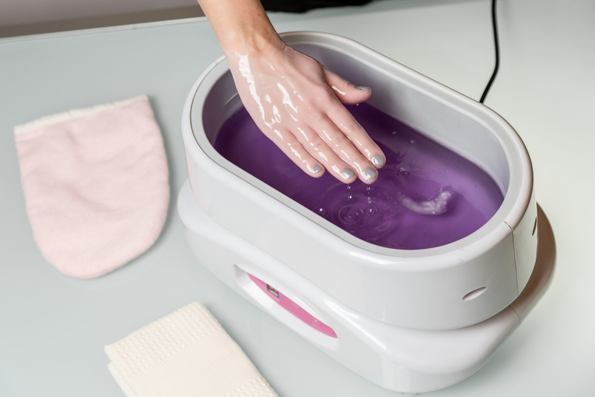Female hands taking procedure in a lilac paraffin wax  bowl. Cosmetological and skincare equipment in a beauty &amp; spa salon.